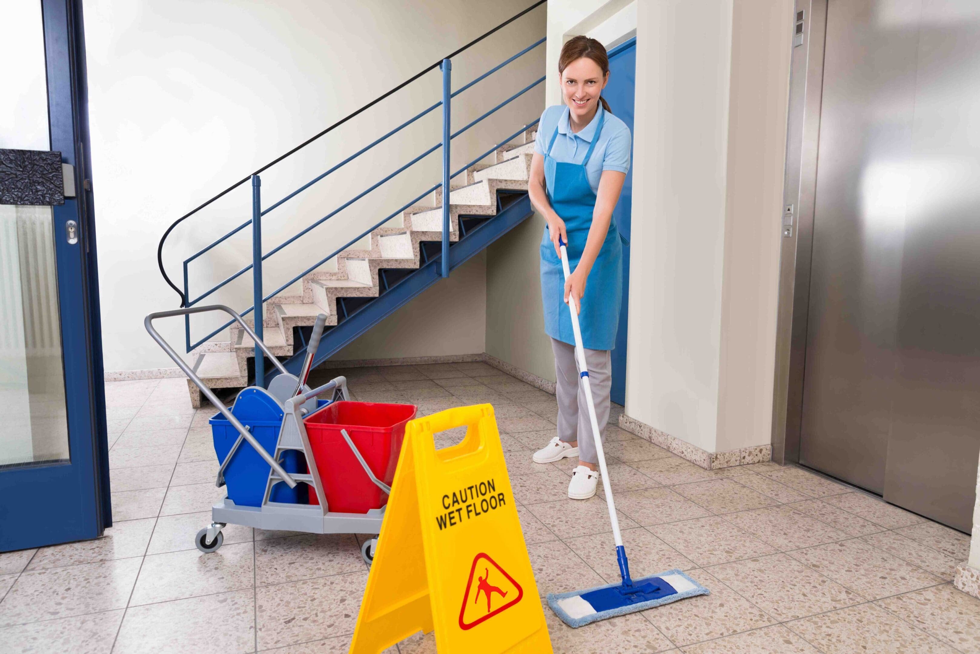 Young Happy Female Worker With Cleaning Equipments And Wet Floor Sign On Floor